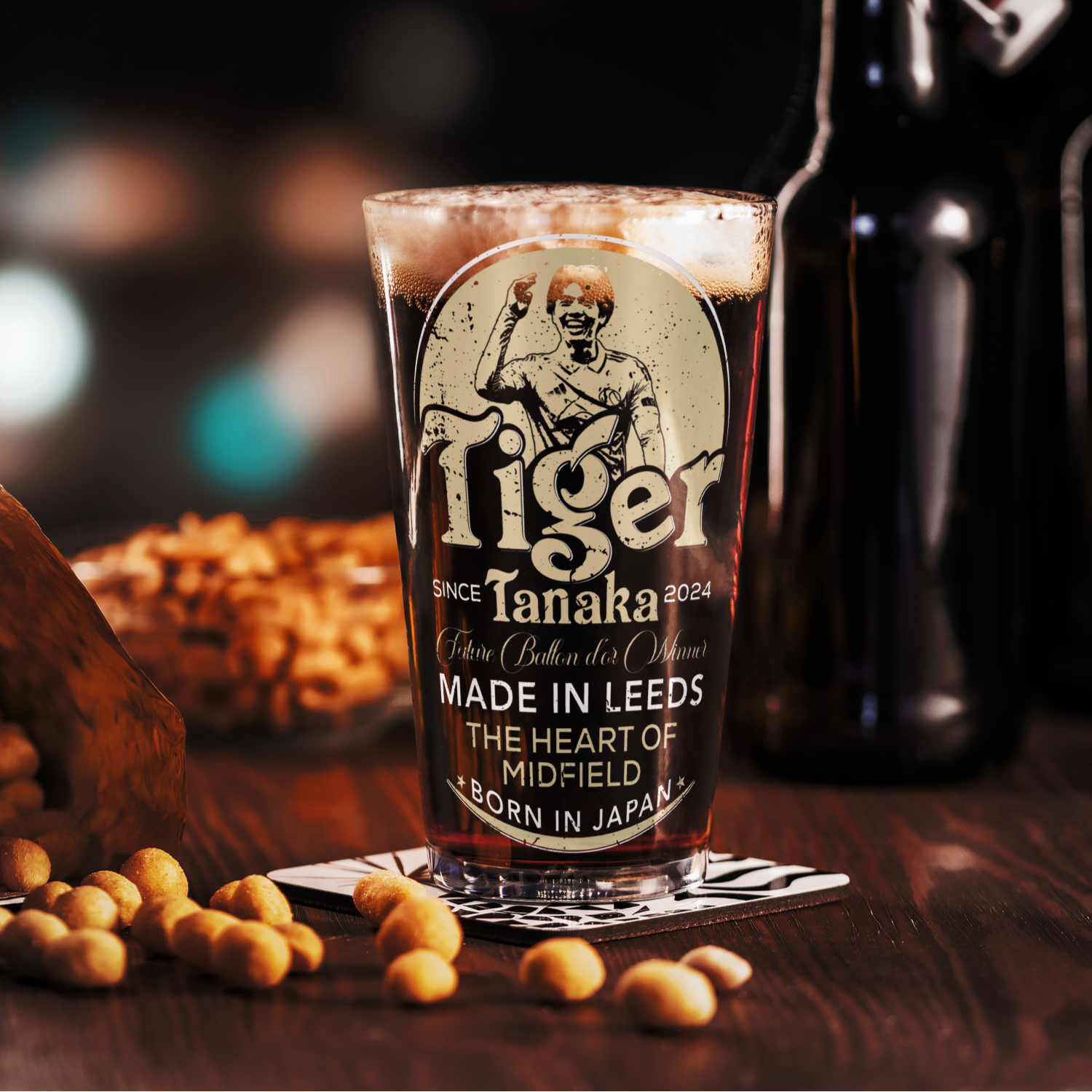 Pint glass with 'Tiger Tanaka' branding on a wooden surface with beer bottles and snacks in the background.